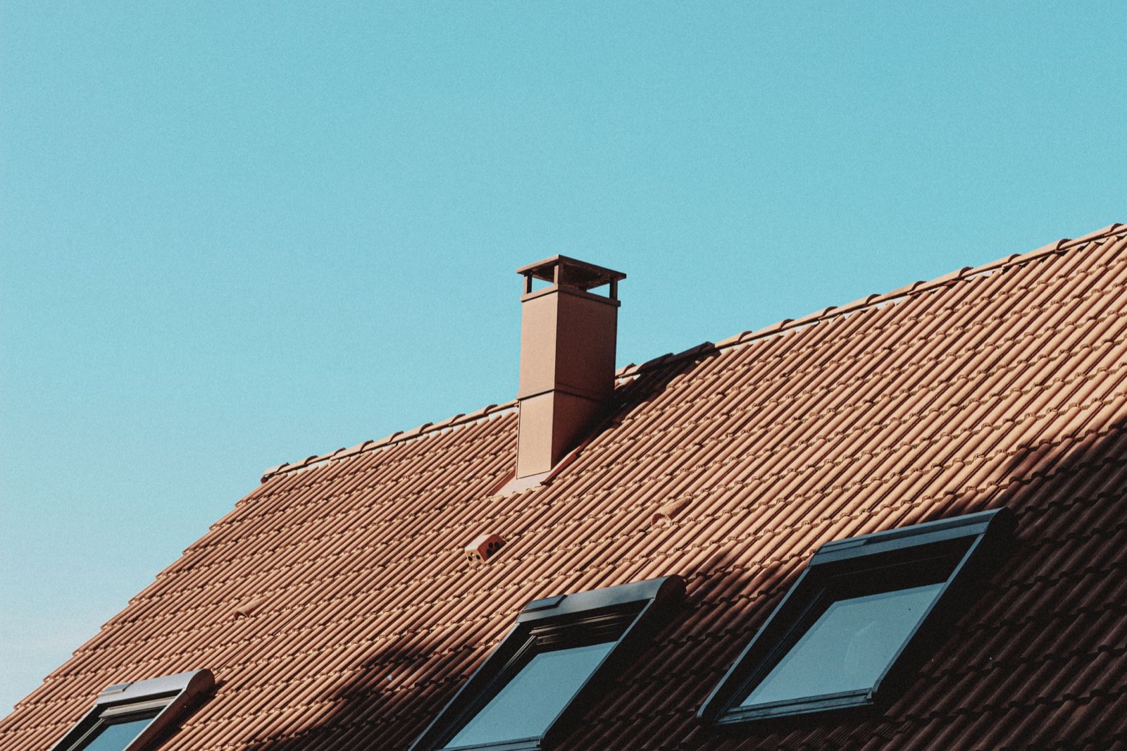 A brick chimney and skylights on the tiled roof of a modern house under a clear blue sky